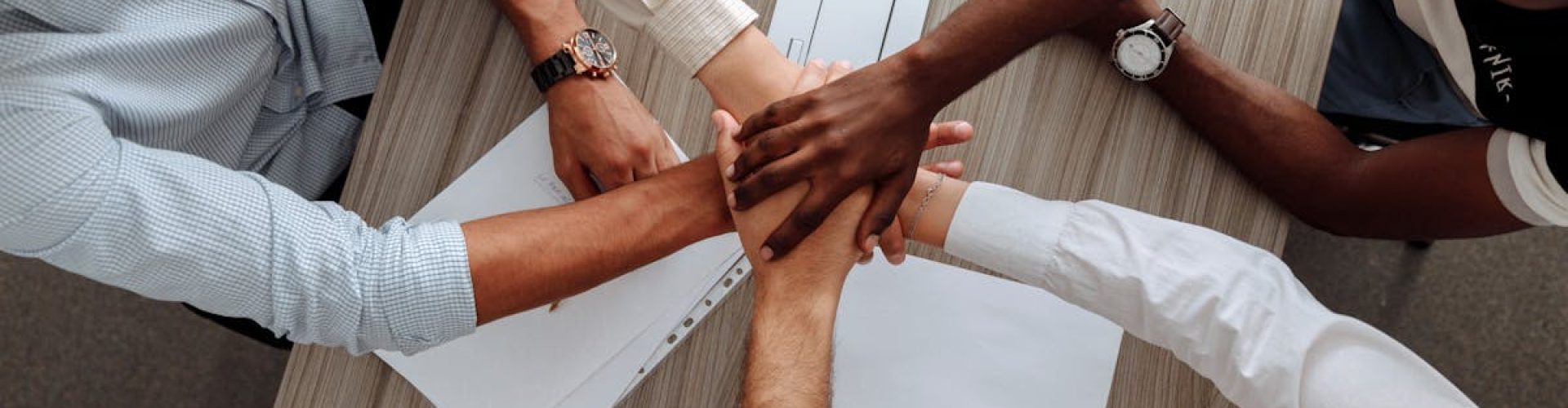 A diverse group of adults join hands in an office, symbolizing teamwork and cooperation.